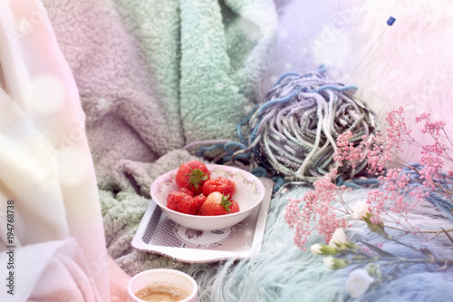 The girl is holding coffee. Fresh strawberries in a plate. On the sofa in the cozy living room. Accessories for hobby knitting. Spring morning. Bouquet of flowers. Card. Free space for text.