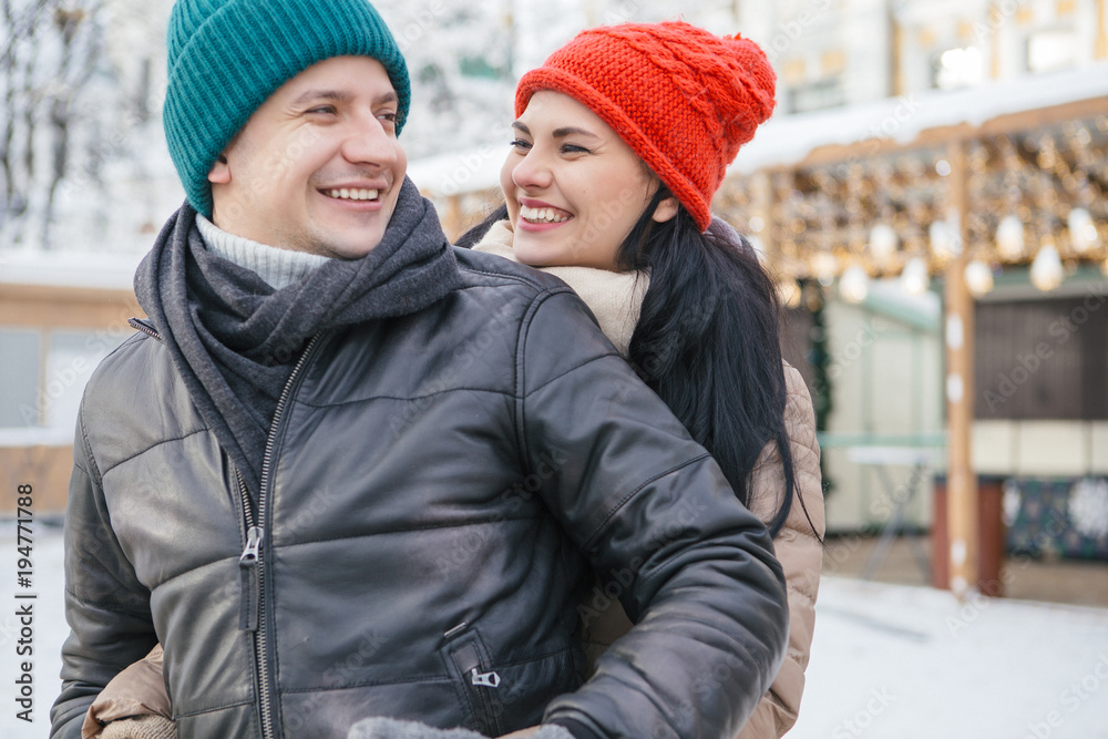 Cheerful smiling caucasian couple of man and woman having fun on european street walk. Casual outfit: jeans, jackets and hats. Winter cold weather with snow. Decorated Christmas tree on a background Cheerful smiling caucasian couple of man and woman having fun on european street walk. Casual outfit: jeans, jackets and hats. Winter cold weather with snow. Decorated Christmas tree on a background