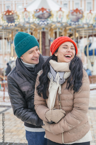 Cheerful smiling caucasian couple of man and woman having fun on european street walk. Casual outfit  jeans  jackets and hats. Winter cold weather with snow. Decorated Christmas tree on a background