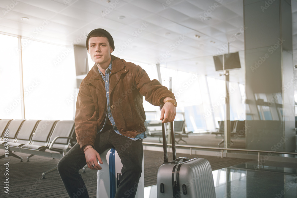 Portrait of weariful young man sitting on luggage while looking in camera in airport. Anticipation concept Portrait of weariful young man sitting on luggage while looking in camera in airport. Anticipation concept