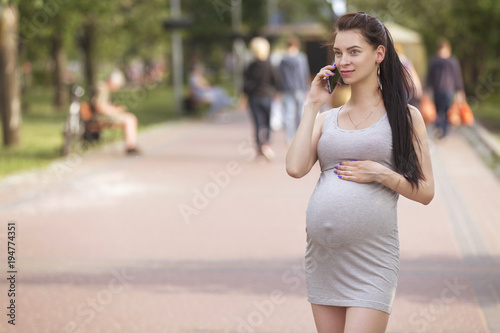 Cute young brunette sexy pregnant woman with long hair in ponytail and in casual dress, walking in european city park, talking on the phone. Sunny warm day, outdoor. Copy space