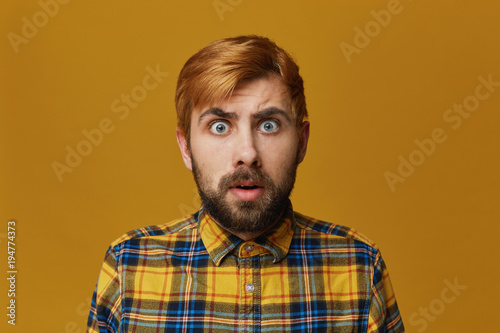 Portrait of stupefied bearded male, with dyed blond gold hair keeps mouth widely opened, wonders about something, isolated over yellow background. Facial expression