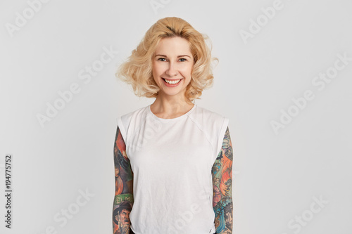 Portrait of young girl wears white t-shirt with pearced nose and tattoo smiling and show her teeth. Facial expression. Isolated over white wall