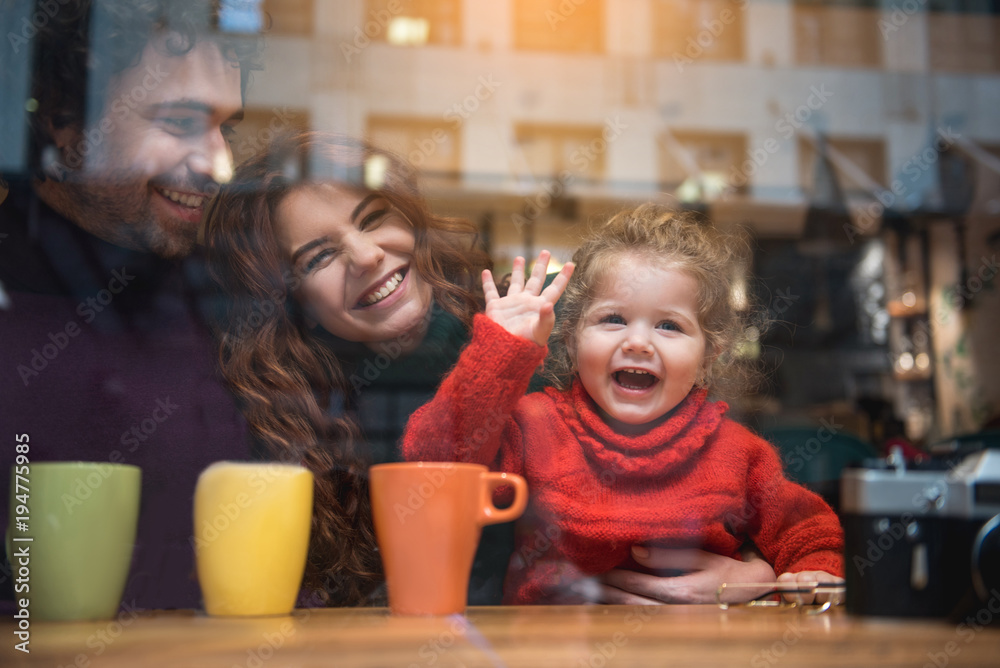 Hello. Portrait of joyful little girl waving hand in front on the window while sitting at table in cafe. Her parents are embracing her and laughing Hello. Portrait of joyful little girl waving hand in front on the window while sitting at table in cafe. Her parents are embracing her and laughing