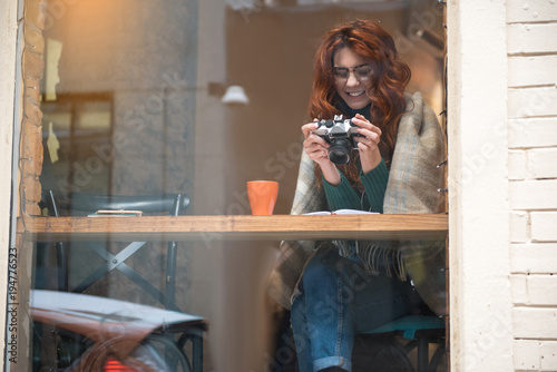 Full length portrait of happy young woman watching interesting photos on camera and smiling. It is visible outside the window how she is sitting in cafe. Copy space