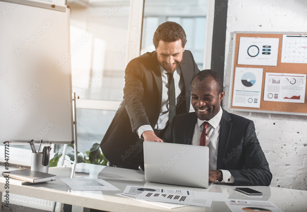 This is good idea. Positive businessman with stubble is leaning over table. He is pointing on screen with smile. Cheerful african manager is sitting at desk with gadget a and expressing gladness This is good idea. Positive businessman with stubble is leaning over table. He is pointing on screen with smile. Cheerful african manager is sitting at desk with gadget a and expressing gladness