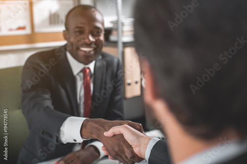 Make deal. Portrait of happy young successful african businessman is wearing official clothes and sitting opposite at table with his partner. He is smiling while shaking hands