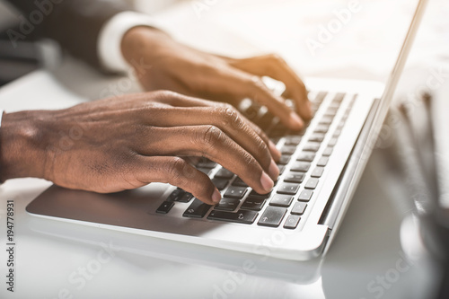 Modern device. Close up of hands of young african businessman is sitting at table and typing on laptop. Selective focus