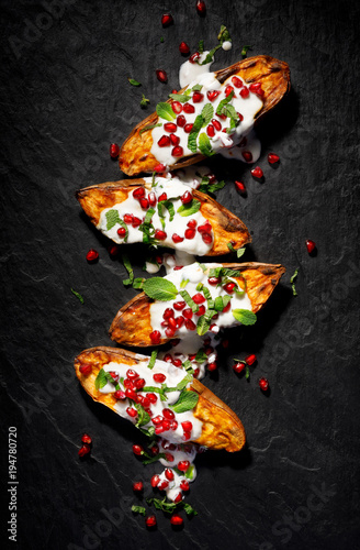 Baked sweet potatoes with garlic mint yogurt sauce sprinkled with pomegranate seeds and fresh mint leaves on a black background, top view