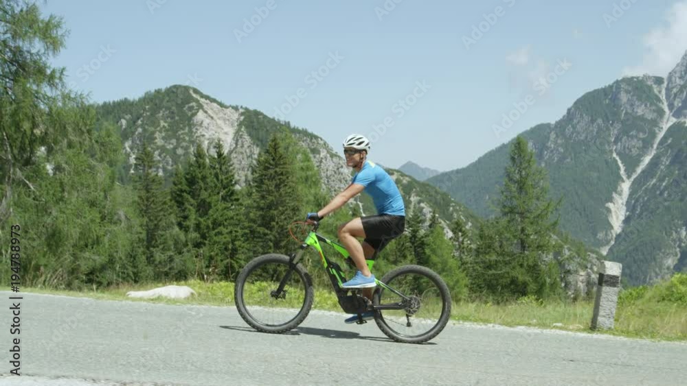 SLOW MOTION CLOSE UP: Cheerful young cyclist riding electric bike easily up a mountain road on sunny summer day. Guy cycling with ease on his e-bike. Fit male rider effortlessly ascending steep road SLOW MOTION CLOSE UP: Cheerful young cyclist riding electric bike easily up a mountain road on sunny summer day. Guy cycling with ease on his e-bike. Fit male rider effortlessly ascending steep road
