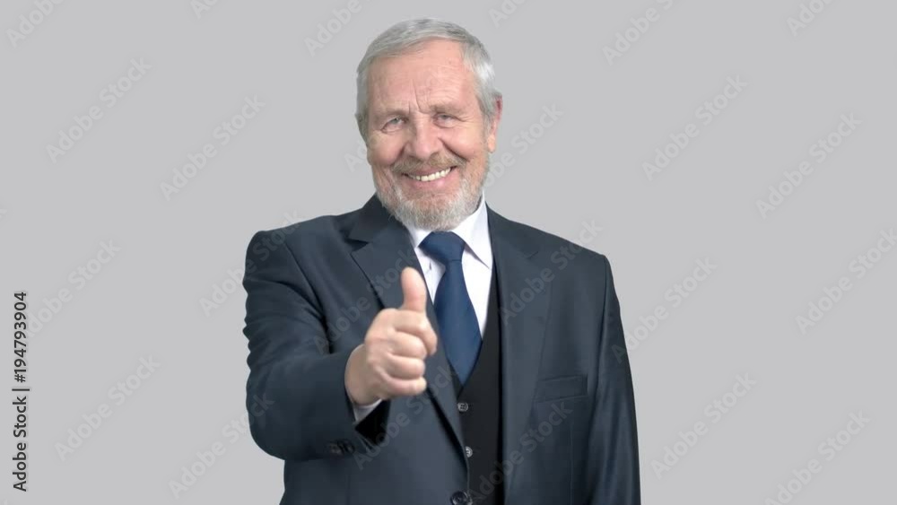 Handsome old businessman gesturing thumb up. Caucasian elderly man in business suit giving thumb up gesture on grey background, portrait. Handsome old businessman gesturing thumb up. Caucasian elderly man in business suit giving thumb up gesture on grey background, portrait.