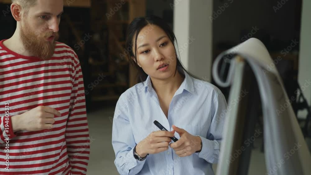 Cheerful asian girl explaining start-up project on flipchart to hipster male colleague at loft office