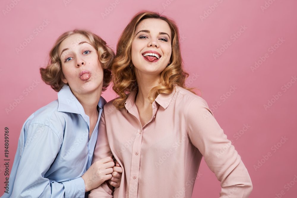 two happy relaxed young girl happily smiling and showing tongue at the camera. Casual solid color shirts and pink background two happy relaxed young girl happily smiling and showing tongue at the camera. Casual solid color shirts and pink background