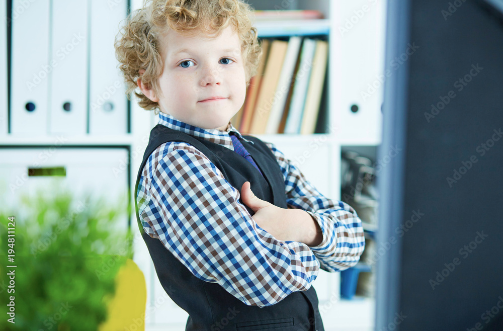 Little Caucasian curly boy presents himself as a businessman posing near the desktop with a monitor in the office Little Caucasian curly boy presents himself as a businessman posing near the desktop with a monitor in the office