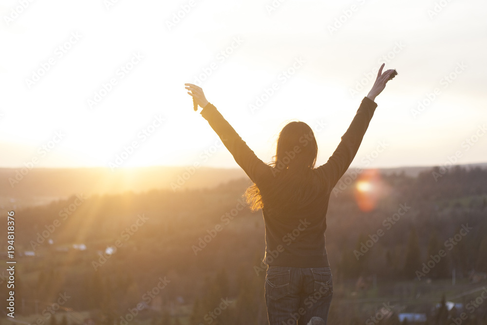 Beautiful black haired young girl watching sunset at rural environment, in the mountains. Raisind her hands in the air