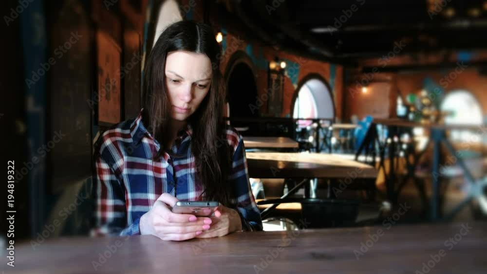 Upset woman sends a message in the phone sitting in a cafe. Dressed in a plaid shirt. Upset woman sends a message in the phone sitting in a cafe. Dressed in a plaid shirt.