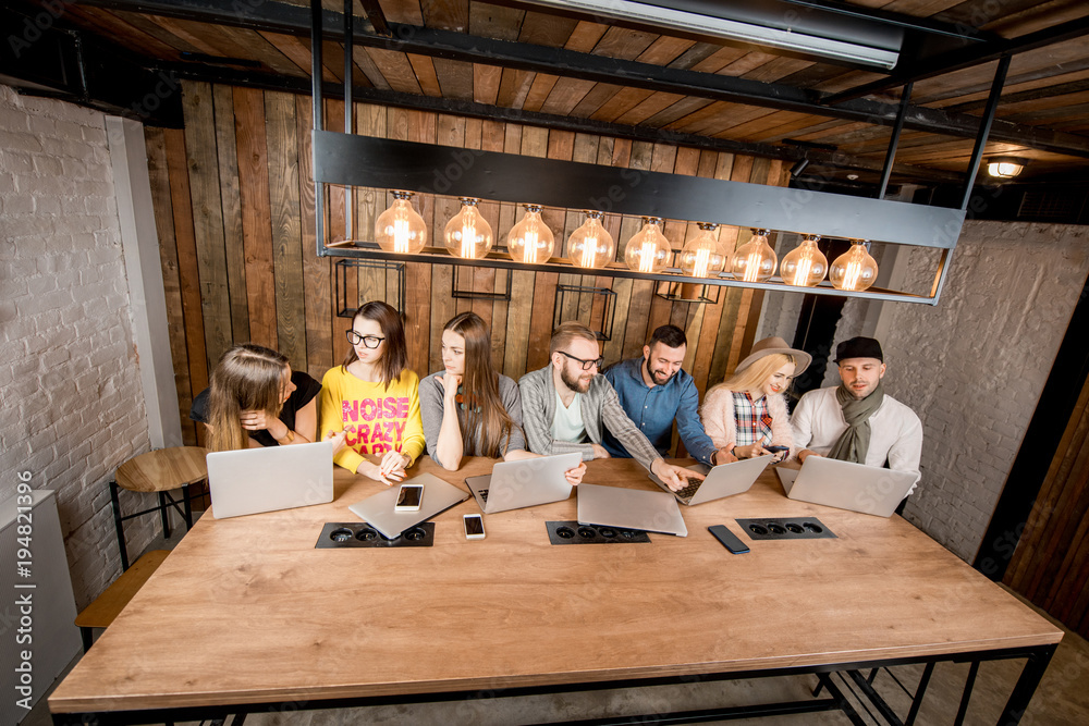 Young coworkers working together with laptops sitting in a row at the wooden table in the modern workspace Young coworkers working together with laptops sitting in a row at the wooden table in the modern workspace