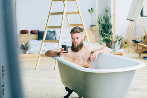 Young caucasian beard man sitting in the bathtub and using smart phone in studio loft interior. Themes for connection, social networking and modern addiction. photo