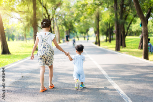 Asian sister hold hands with small children walking on the road in park with rays of sunlight..