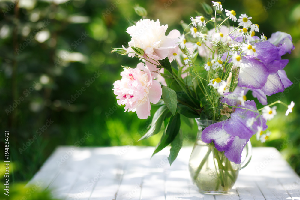 Bouquet of white peonies, chamomiles and iris flowers in glass vase. Summer background