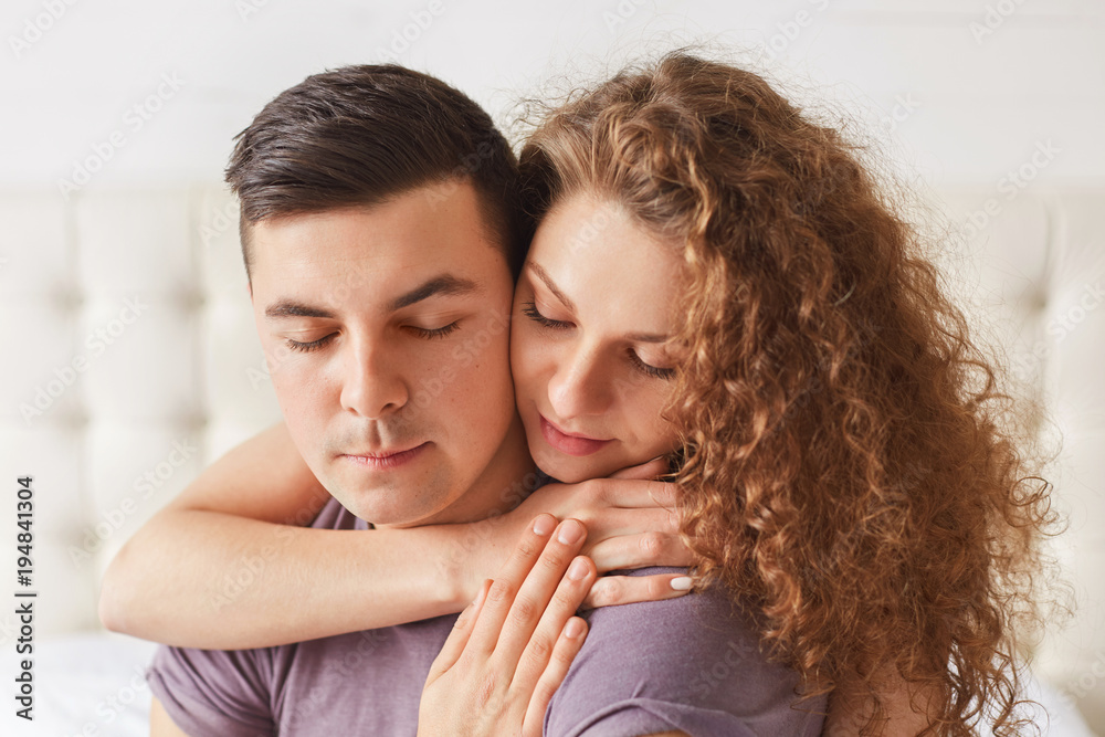 Close up shot of delighted couple enjoy togetherness. Lovely young woman with curly hair embraces her boyfriend, keep eyes shut, feel support and love to each other. Mutual relationships concept