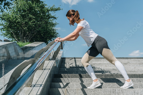 Sunny summer day. Young woman in sportswear doing stretching exercises outdoor. Girl doing warm-up on steps before training. Exercise in street, sports exercises, workout. On background blue sky.