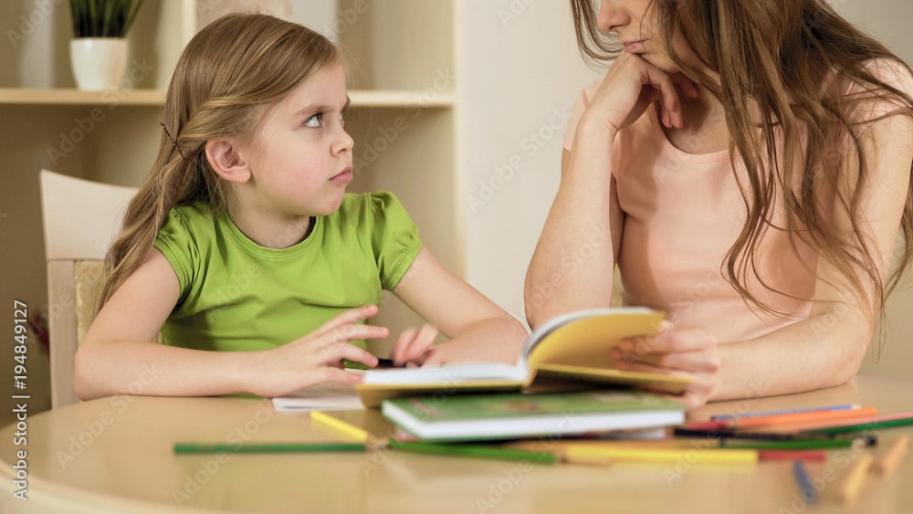 Upset little girl with misunderstanding looking at her mother doing school tasks Upset little girl with misunderstanding looking at her mother doing school tasks