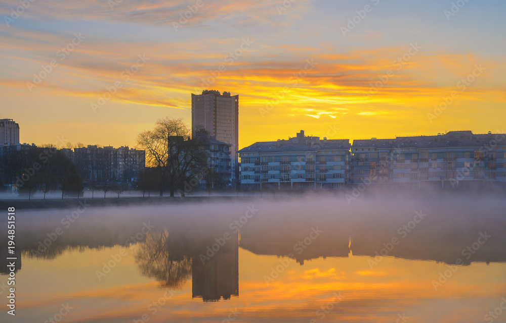 Landscape in city park. Heavy fog on water in morning. Multistory buildings on river bank