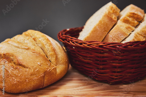 Fresh bread made from wheat flour in a wicker basket made of dark wood lies on a wooden board and against a dark background. Lighting from above through the hole. Slices of freshly baked bread.