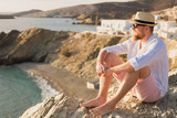 Bearded romantic male traveler in a hat and glasses meets the dawn on the shore of the bay