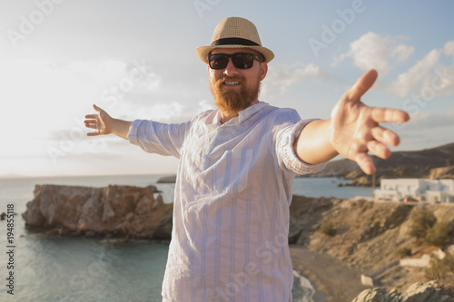 Red-haired bearded hipster traveler man with open hands is standing with his back to the sea and smiling in the rays of the morning sun