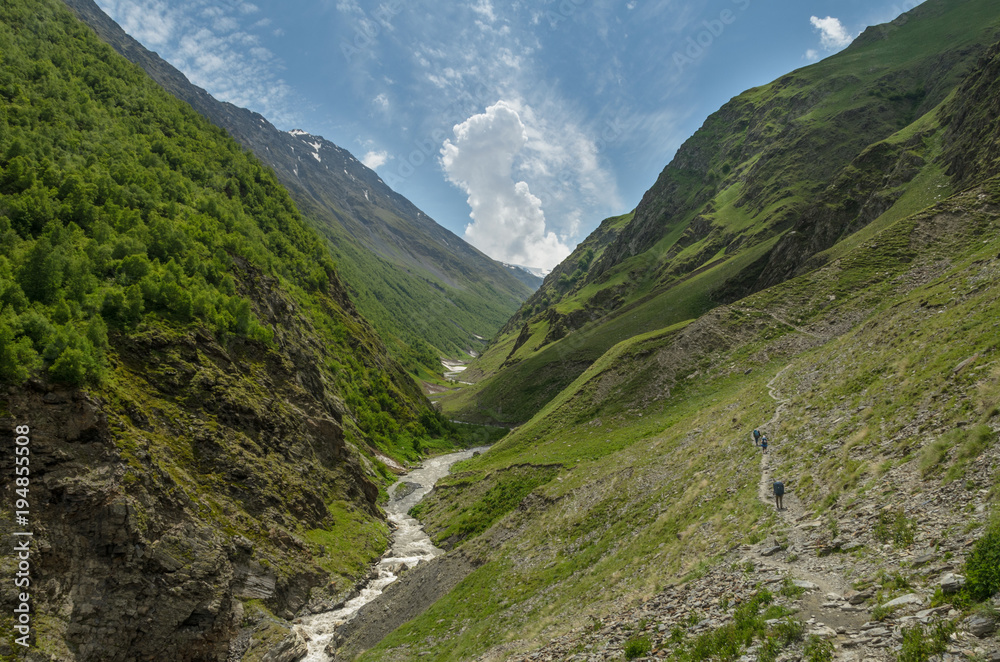 Three backpackers walking along the trail along the mountain river valley. Hills covered with green grass. A couple of clouds in the blue sky. Hot summer day at Caucasus mountains at Tusheti, Georgia Three backpackers walking along the trail along the mountain river valley. Hills covered with green grass. A couple of clouds in the blue sky. Hot summer day at Caucasus mountains at Tusheti, Georgia