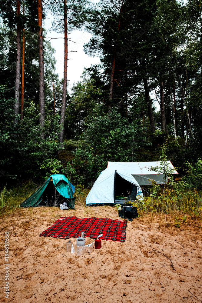 Two tents, a tarp and a blanket at a beach in Sweden with gas stove coffee cup and camping gear. Trees and forest in the background.