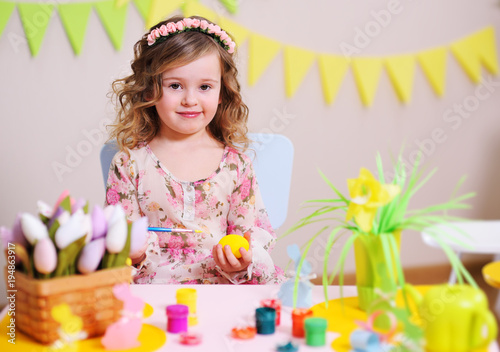 girl in a smart dress paints an egg sitting at a table with an Easter decoration