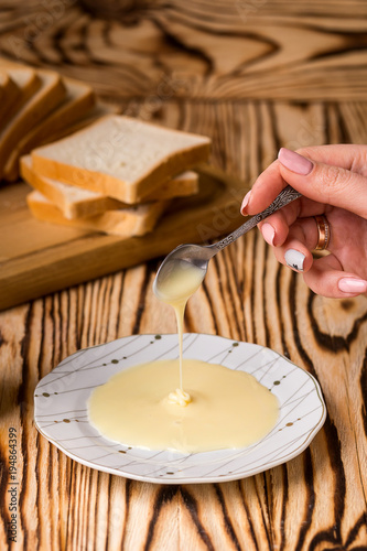 A bowl and a bottle of condensed milk and a teaspoon on a wooden table with background of toasts and tea cup