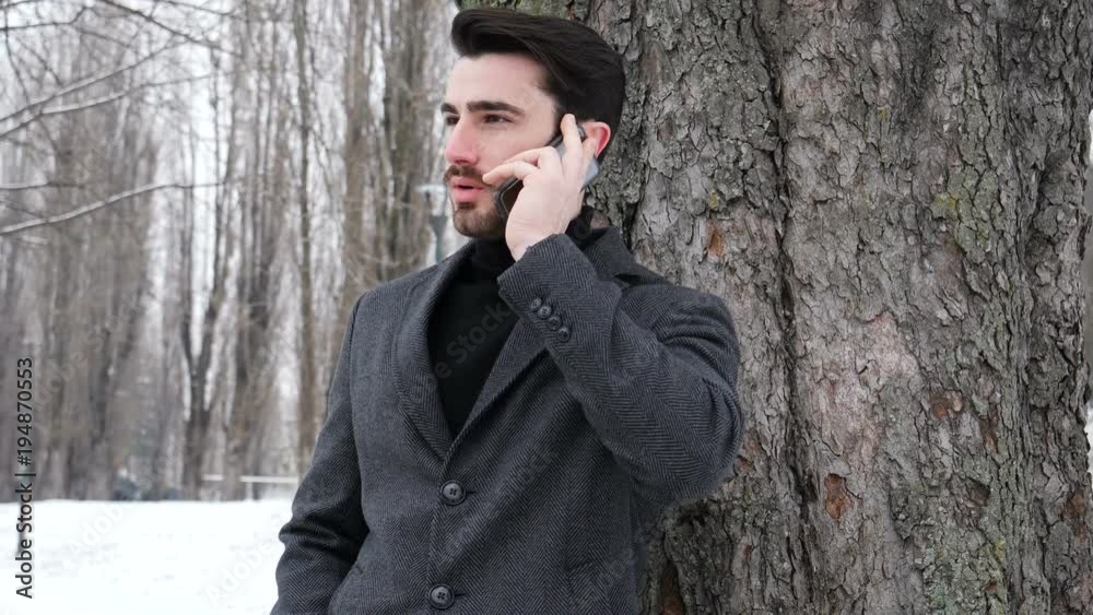 Handsome trendy man wearing black jacket standing and talking on cell phone, outdoor in city setting in winter day shot
