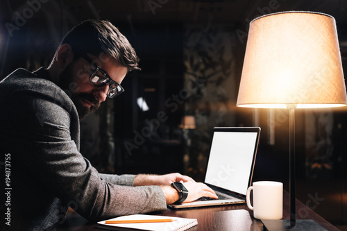 Bearded young coworker wearing glasses typing text on contemporary laptop in modern loft office at night. Business man sits by the wooden table with lamp working on new startup project. Film effect