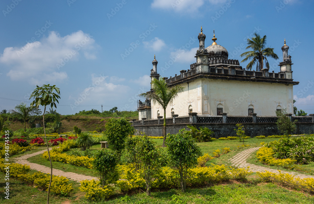 Madikeri, India - October 31, 2013: White and gray Royal mausoleum set in green garden of domain Raja Tombs under blue sky with clouds. Bushes and trees.