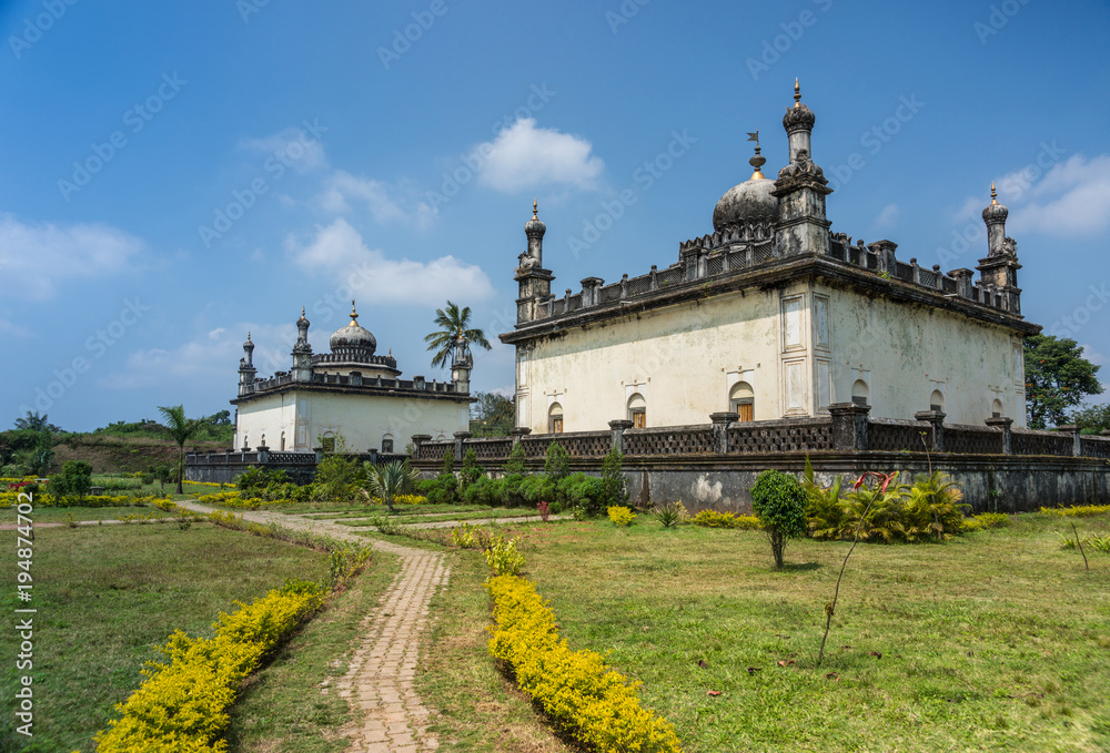 Madikeri, India - October 31, 2013: Two largest white and gray Royal mausoleum set in green garden of domain Raja Tombs under blue sky with clouds. Bushes and trees.
