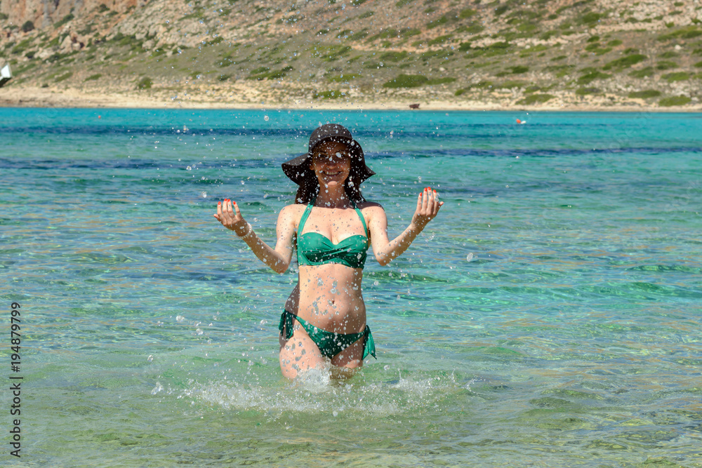 Portrait of a cheerful young girl in a black hat and green swimsuit standing in the emerald sea water and playing with water, making splashes, copy space