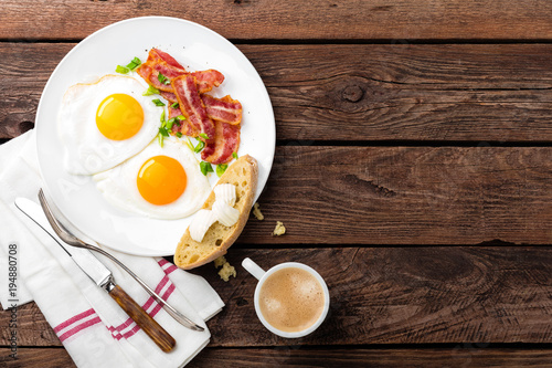 Fried eggs, bacon and italian ciabatta bread on white plate. Cup of coffee. Breakfast. Top view. Wooden background