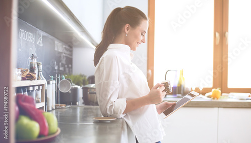 Young woman using tablet in kitchen at home and drinking coffee