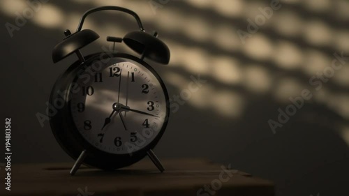 Alarm clock on night table in bedroom ticking time in early morning with sunlight and shadows on the wall in background