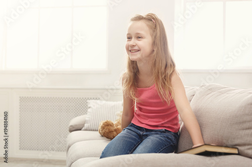 Happy little female child and her teddy bear reading book on sofa at home