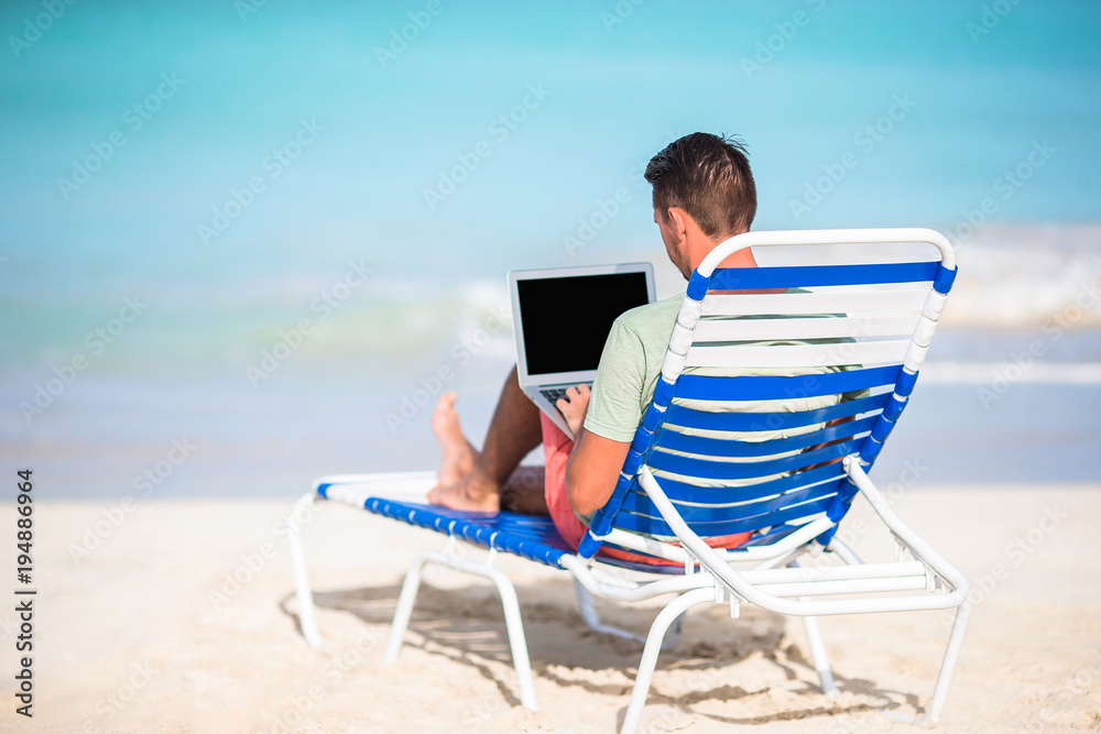 Young man with laptop on tropical beach. Man sitting on the chaise-lounge with computer