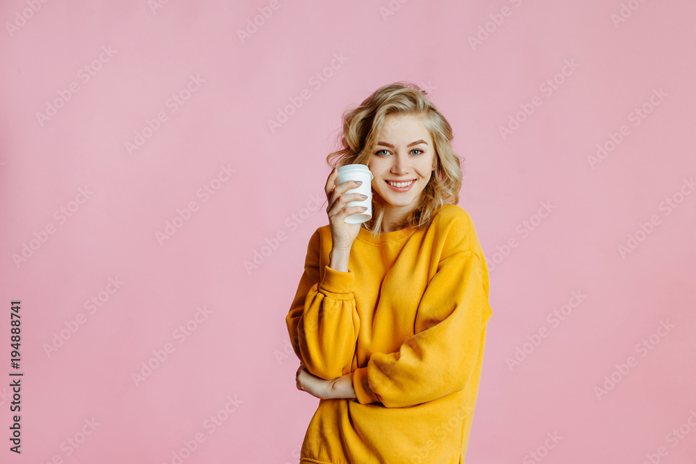 close-up portrait of cheerful young caucasian female with curly blonde hair, in yellow sweater poses on a pink background. woman holds in hand a glass with coffee and smiles close-up portrait of cheerful young caucasian female with curly blonde hair, in yellow sweater poses on a pink background. woman holds in hand a glass with coffee and smiles