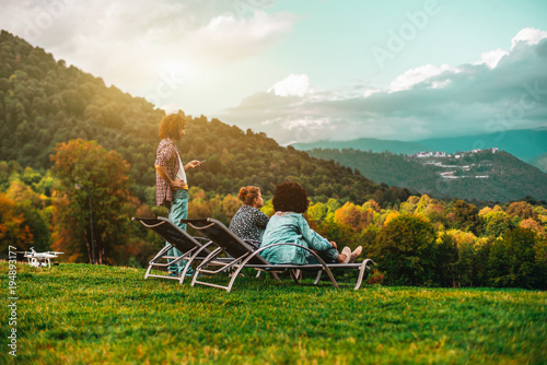 Group of three friends: two girls are chilling on daybeds on the lawn and one bearded man standing next to them and using his smartphone; drone on the ground near, colorful beautiful autumn landscape