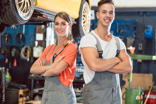 Portrait of two skilled auto mechanics looking at camera with confidence while wearing gray overall in a modern automobile repair shop with experienced employees