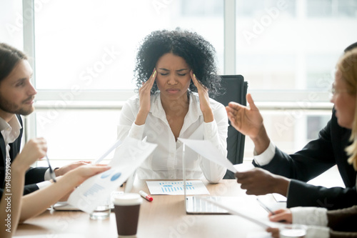Stressed overwhelmed african businesswoman feels tired at corporate meeting, exhausted black female boss suffering from headache touching temples at team briefing, stress at work or migraine concept