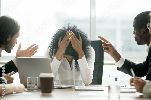 Upset depressed black woman leader suffering from gender discrimination inequality at work, diverse men colleagues pointing fingers scolding bullying frustrated african businesswoman at workplace